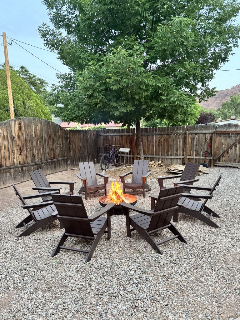 Outdoor backyard seating area with eight black Adirondack chairs arranged in a circle around a fire pit with flames, gravel ground, a large leafy tree, a wooden fence, a bicycle, and some stacked wood in the background.