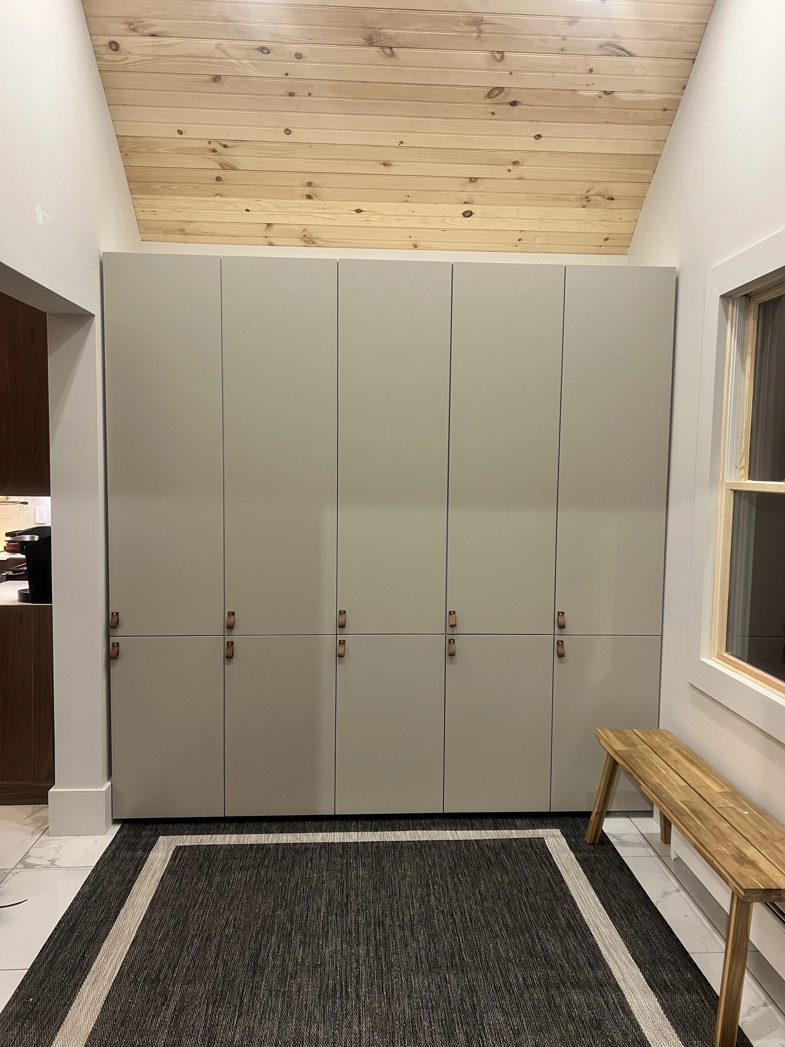 A room with tall, light green cabinets with brown handles, a rug, a bench, a window on the right, and a wooden ceiling.