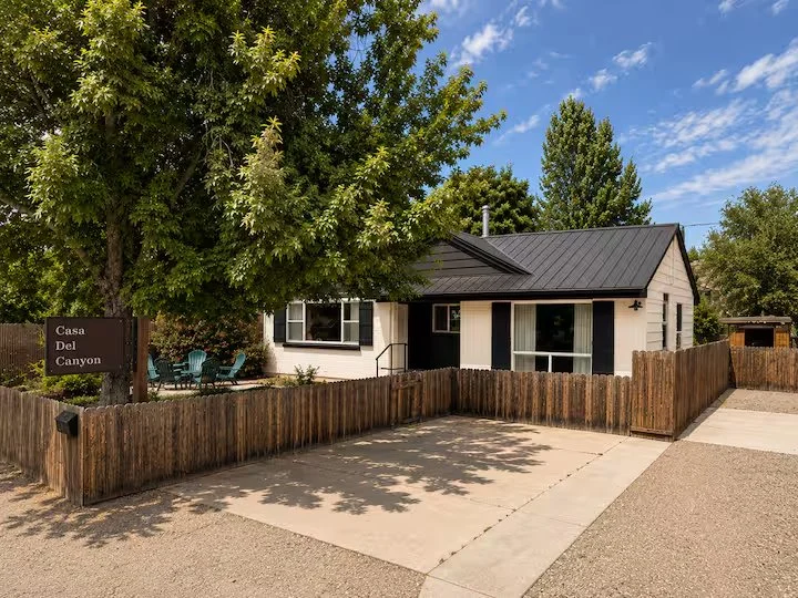 A small house with a metal roof, surrounded by a wooden fence, with a large green tree in front and a bright blue sky overhead.