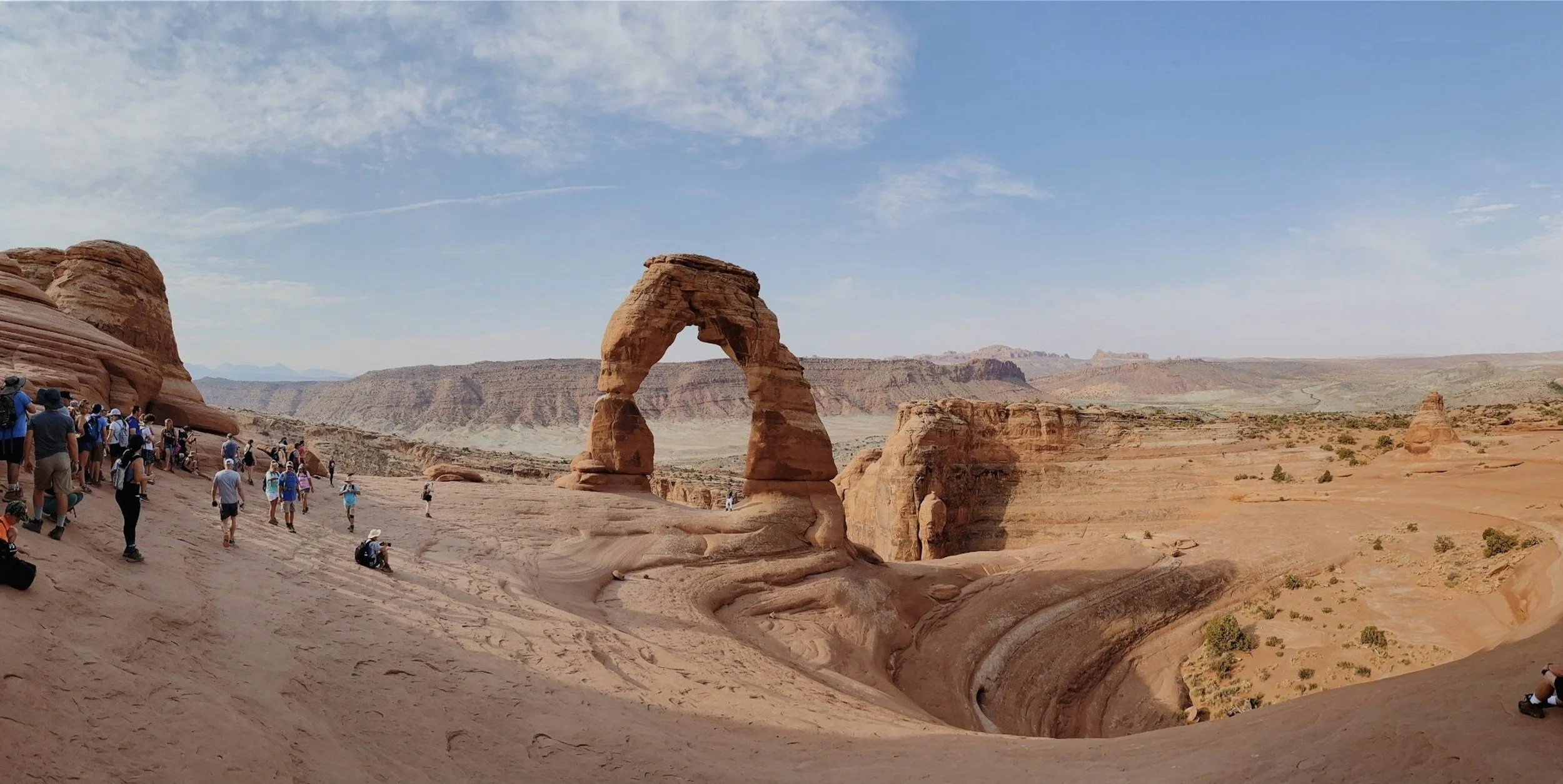 Tourists walking and sitting on sandstone formations near Delicate Arch in Arches National Park, Utah, with a clear blue sky and distant landscape.