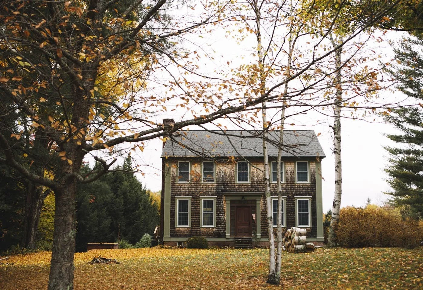 A two-story house with brown wooden shingles, surrounded by trees with autumn leaves, some fallen on the lawn.