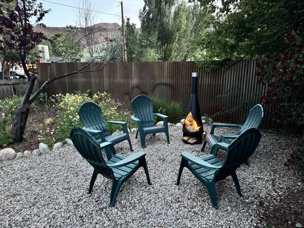 Five teal plastic outdoor chairs arranged in a circle around a chiminea with a small fire, on a gravel patio in a backyard with trees and bushes.