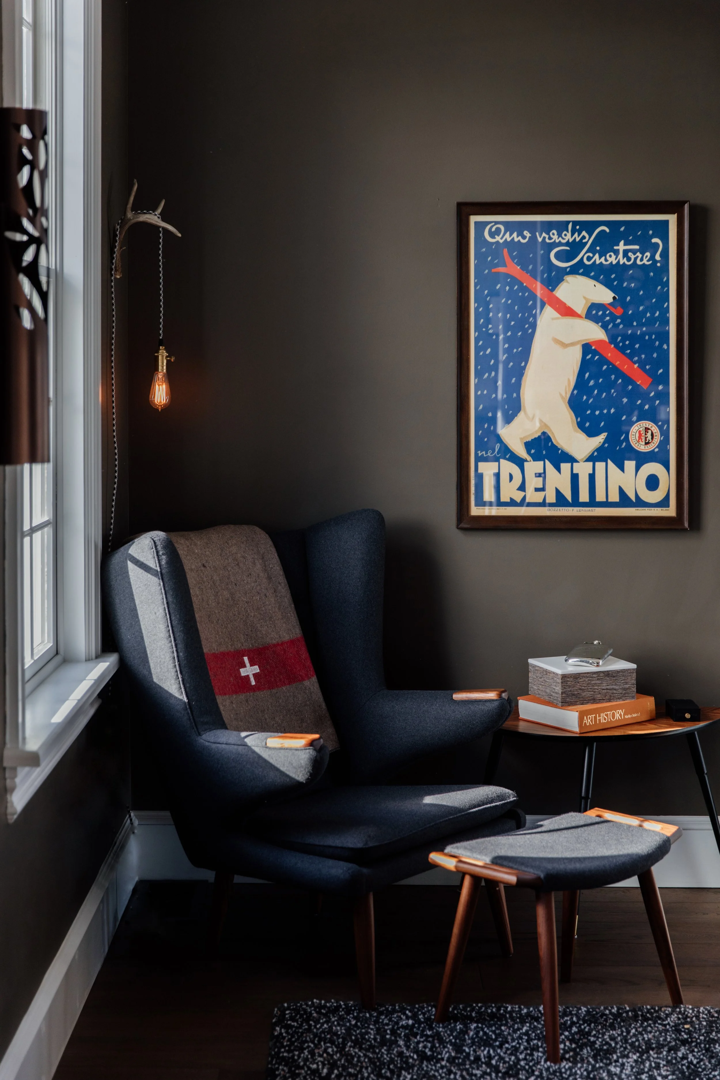 A cozy reading nook with a dark gray wall and modern furniture. There is a pair of high-back armchairs, one with a Swiss flag-themed blanket, a small round table with books, a box, and a remote. A framed poster of a bear holding a red pole with text "Who dares, wins" and "trentino" hangs on the wall. A window with white trim is on the left, and a hanging Edison-style light bulb is beside the chairs.