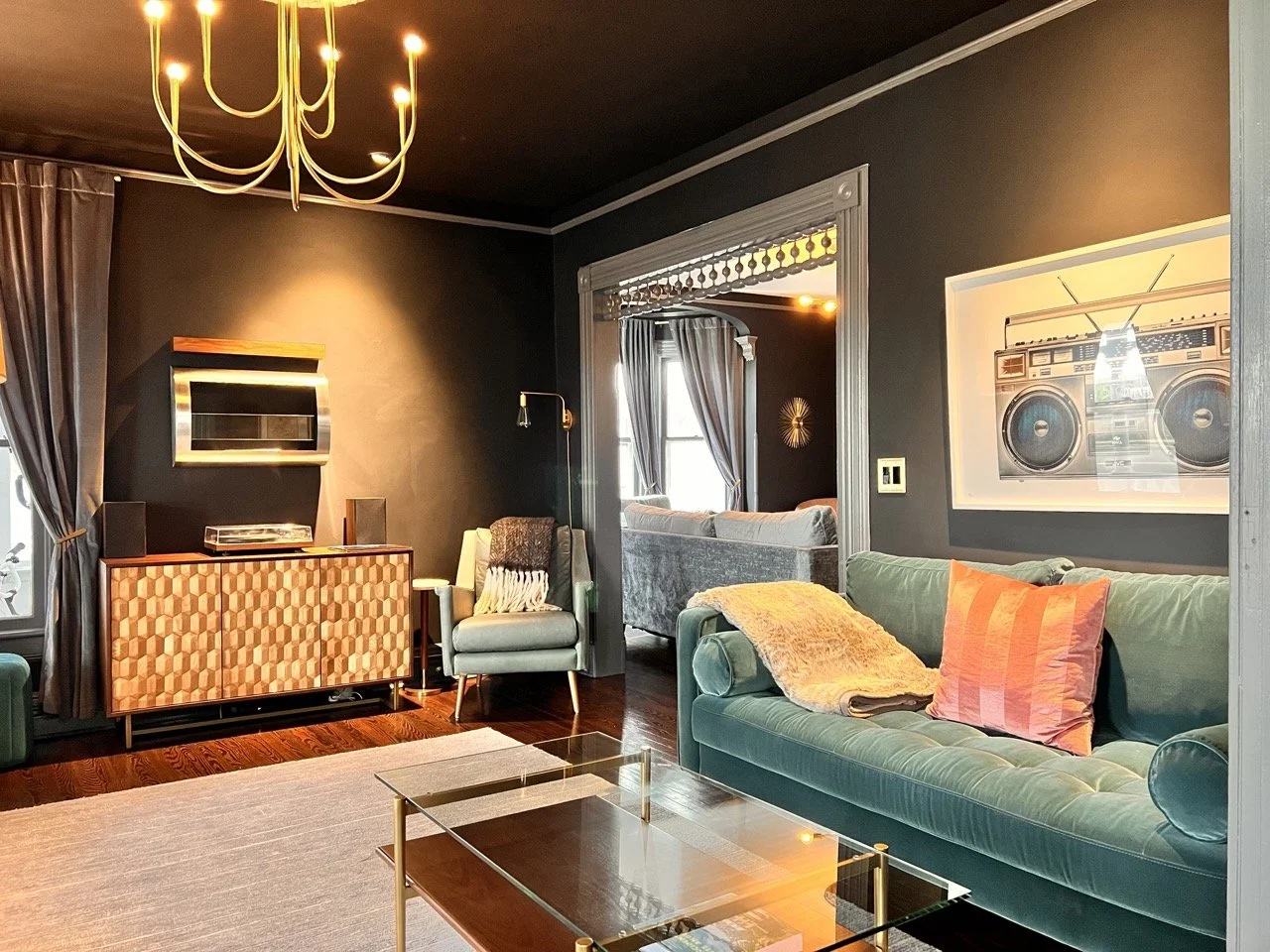 Living room with a teal velvet sofa, a pink pillow, a beige throw blanket, a glass coffee table, a patterned cabinet, and a black wall with a framed boombox photograph. There's a window with gray curtains and a modern chandelier hanging from the ceiling.