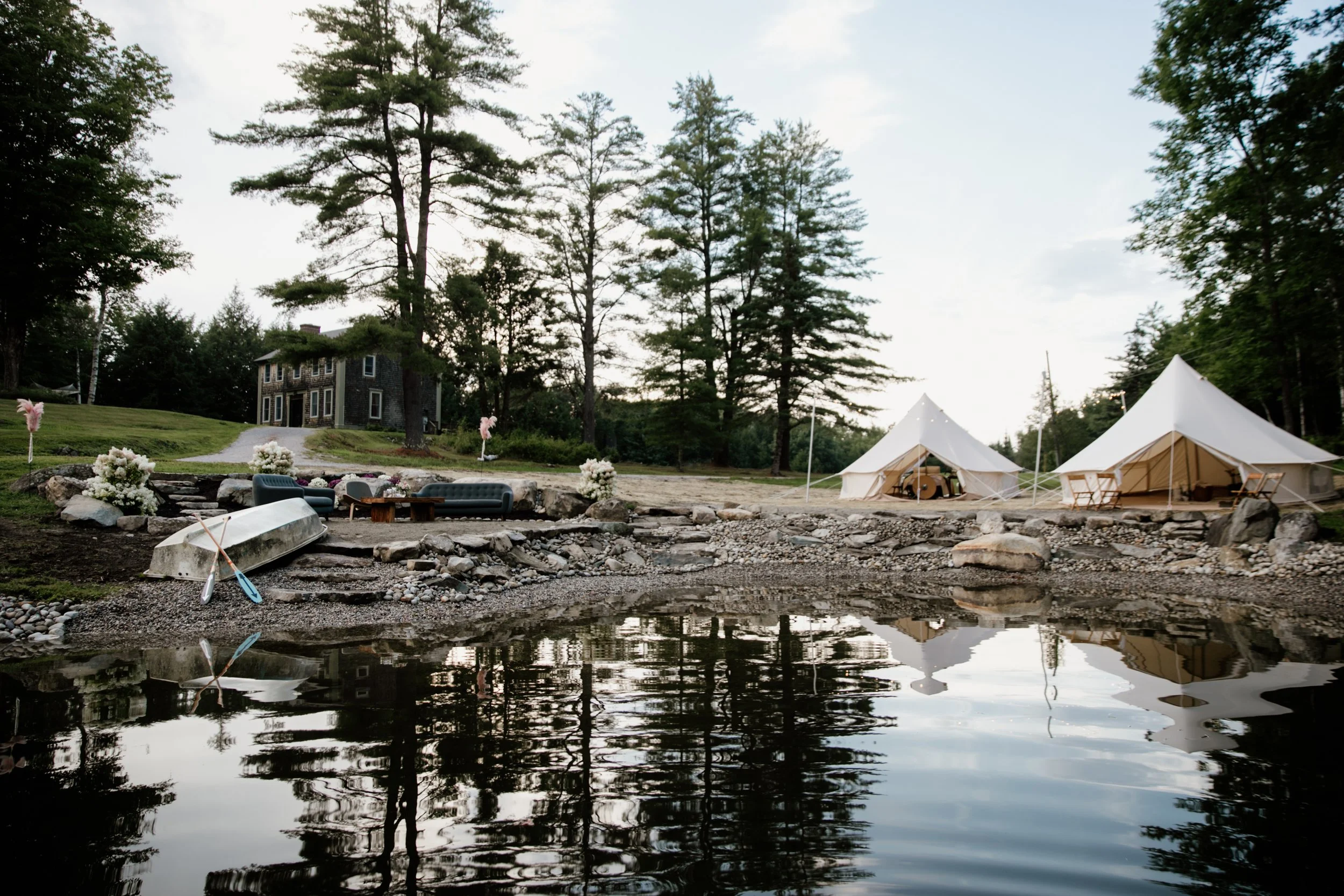 A scenic outdoor setting with two white tents on a rocky shore near a body of water, surrounded by tall trees and a house in the background, decorated with pink bows and flower arrangements.