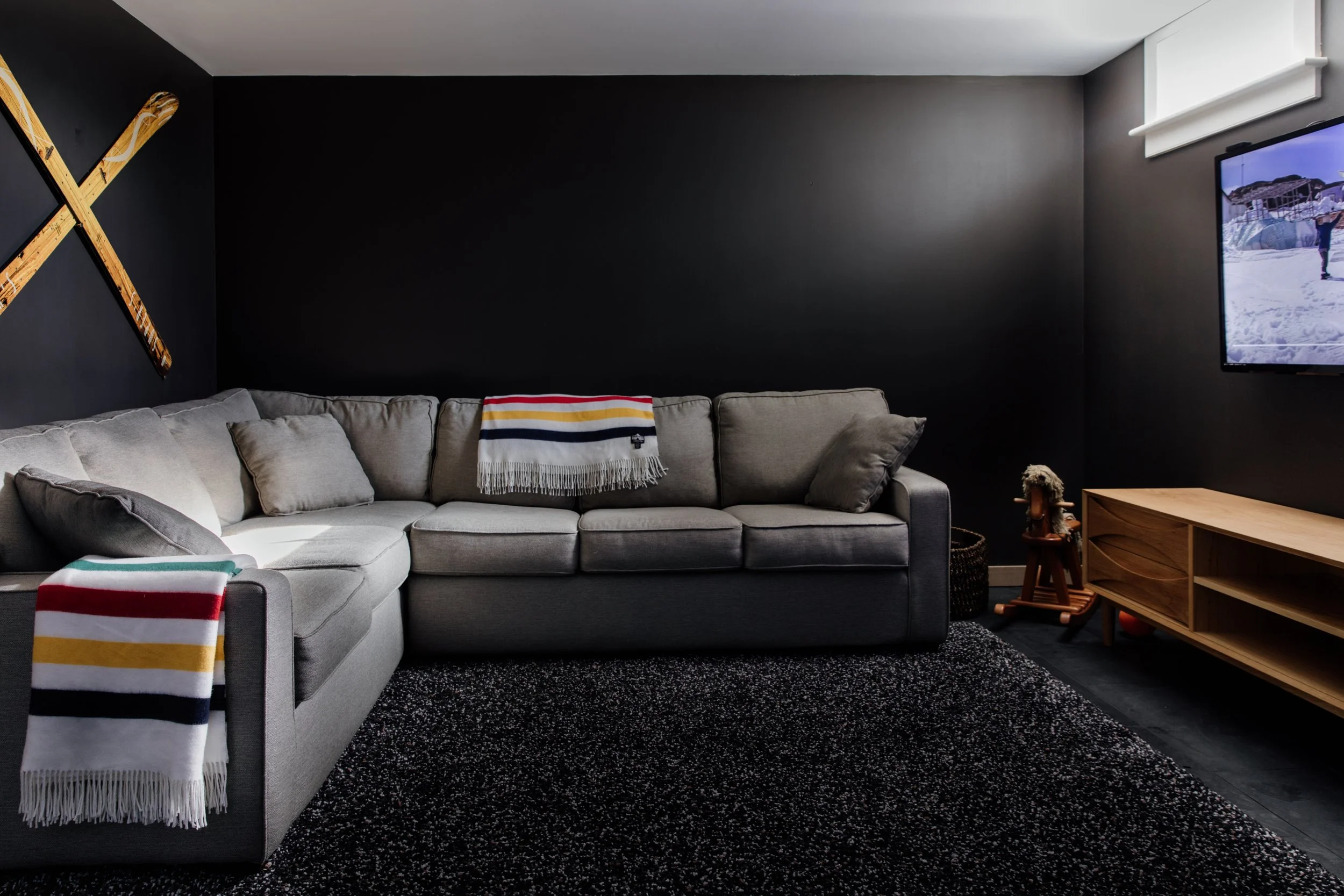 Living room with a light gray sectional sofa, black wall, two colorful striped throws, wooden skis on the wall, a television, and a wooden entertainment center.