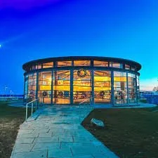 A modern round glass building with multiple floors, illuminated from inside, situated in an open outdoor area during dusk.