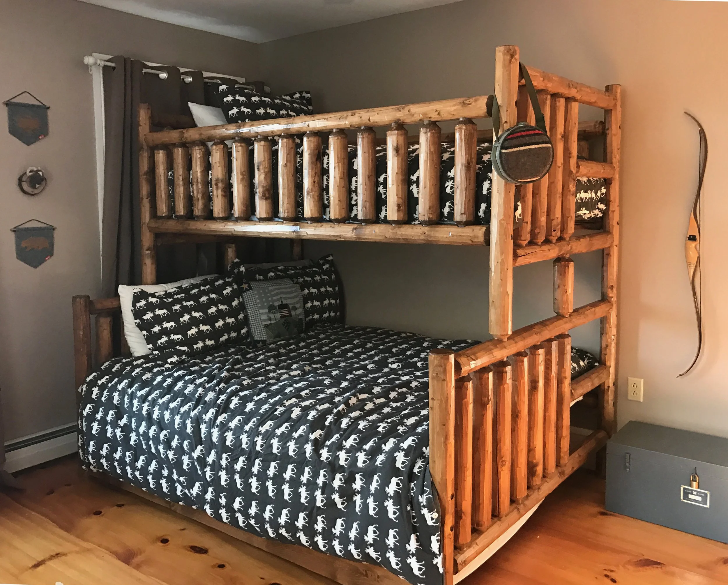 A bedroom with a wooden bunk bed, featuring a double bed on the bottom and a twin bed on top. The bedding has a black and white dinosaur pattern. There are dark gray curtains, a wall hanging, and a decorative item attached to the wall. A gray storage chest is on the floor, and a bow hangs on the wall.