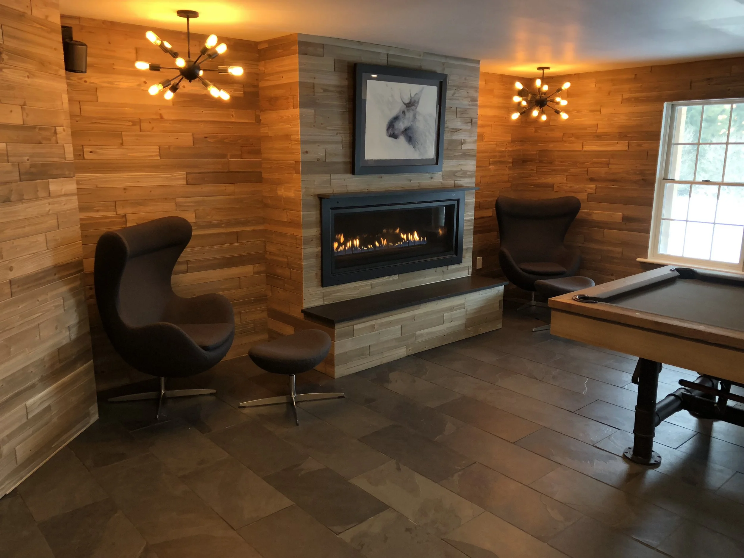 Living room with wooden wall paneling, a modern gas fireplace, two black lounge chairs with round matching ottomans, a window with white trim, and a pool table.