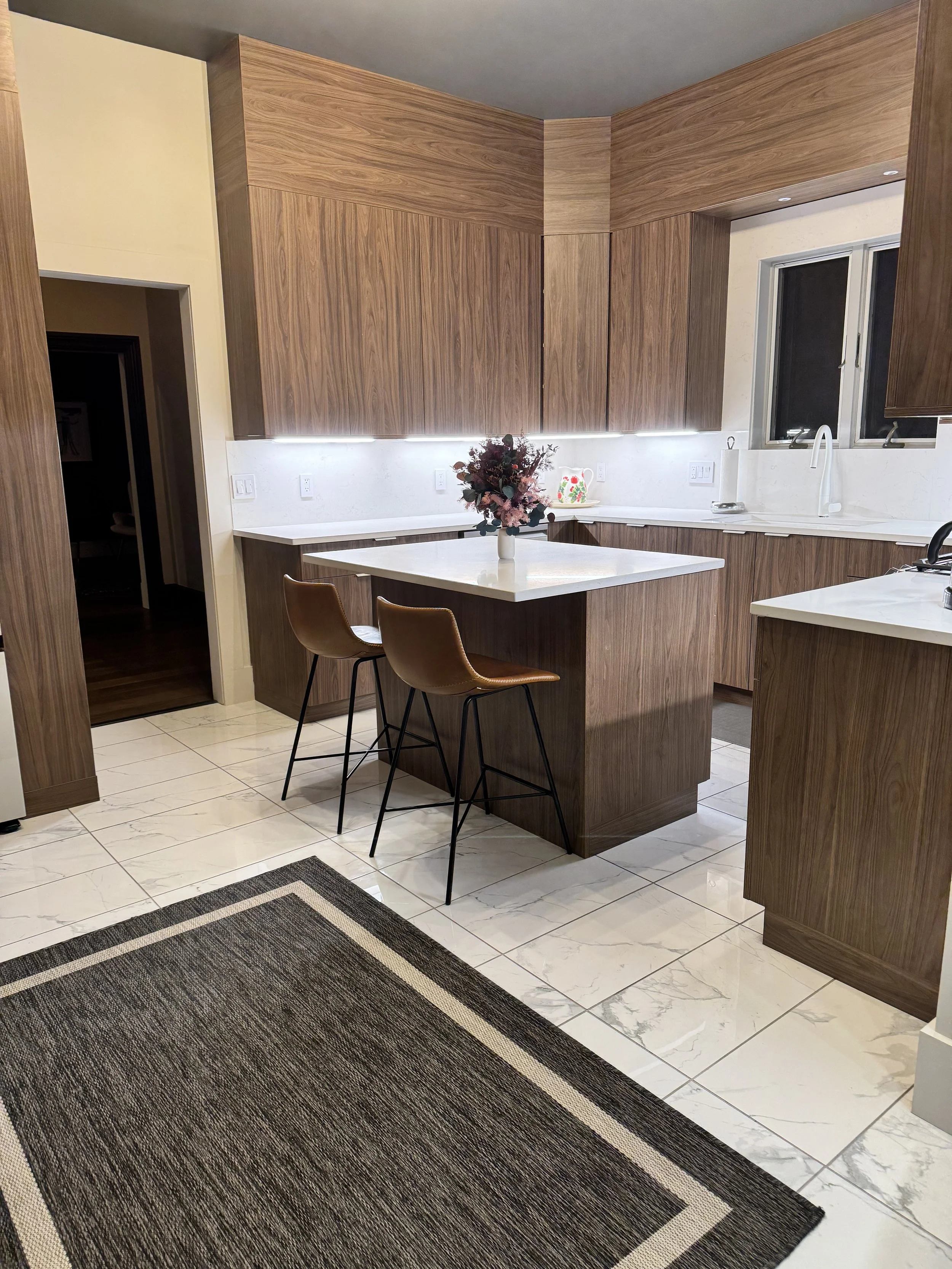 A modern kitchen with wooden cabinets, a white countertop, and a kitchen island with a white vase and flowers. There are two brown chairs at the island, a window above the sink, marble-patterned floor tiles, and a dark rug in the foreground.