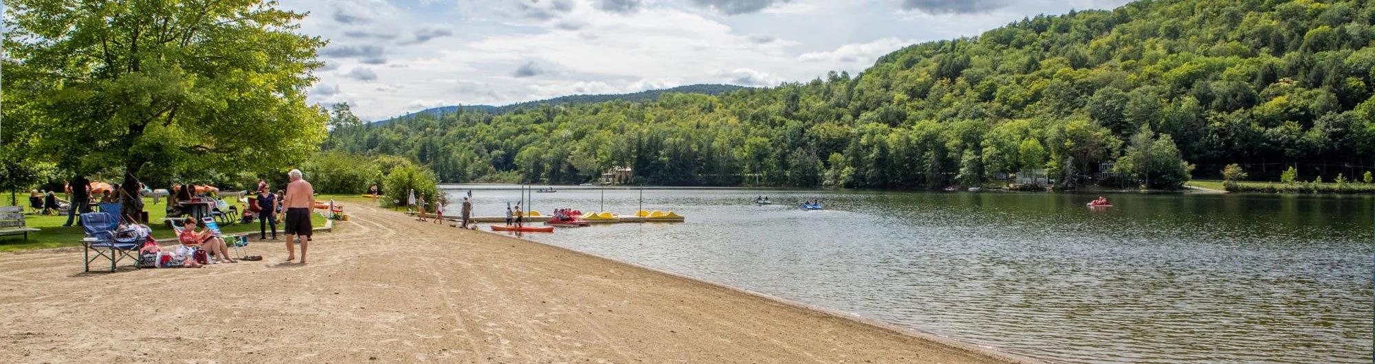 People on a beach by a lake with boats and kayaks, surrounded by green trees and hills under a partly cloudy sky.