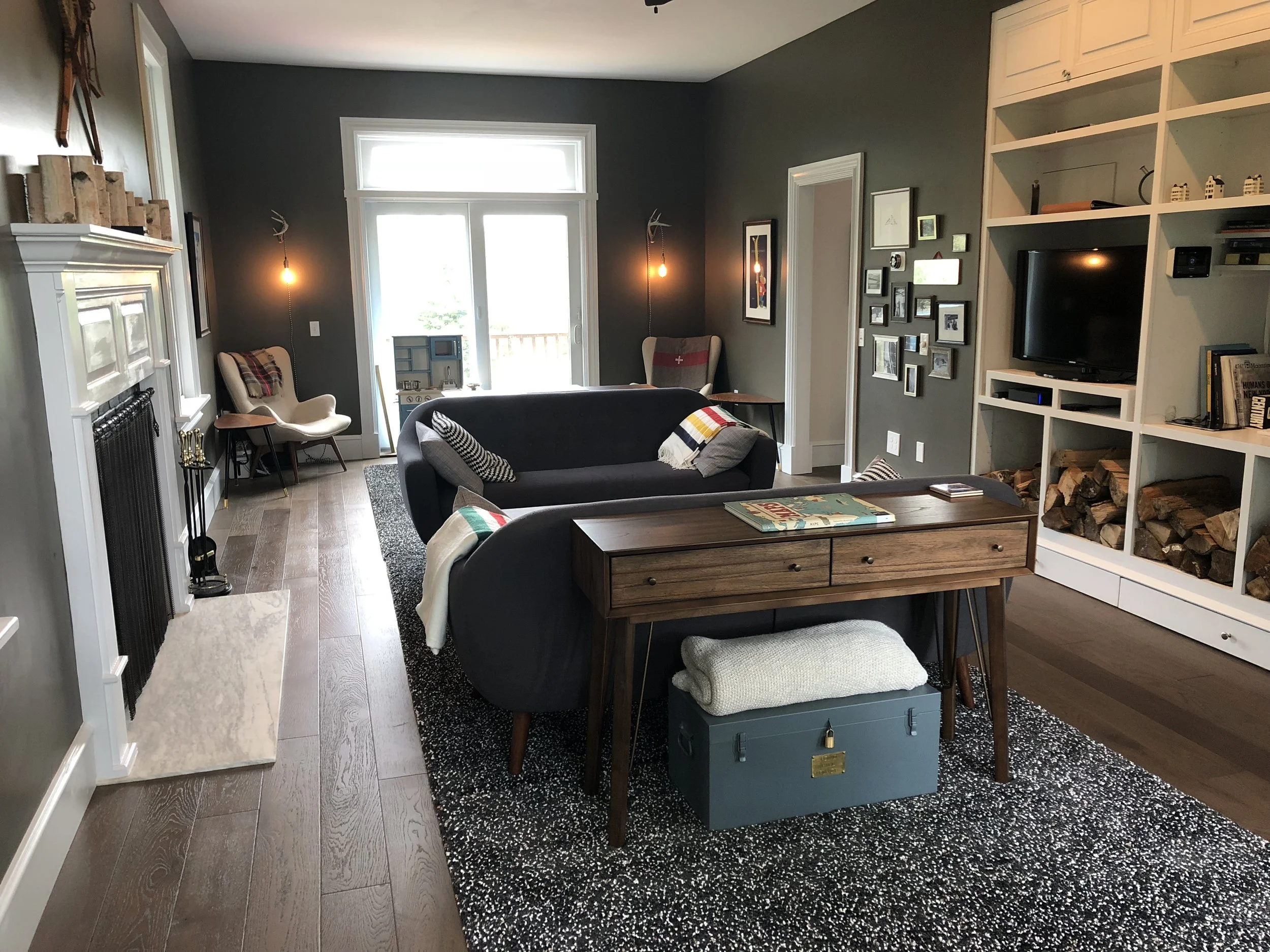 Living room with dark green walls, wood flooring, white fireplace, black sofa, wooden coffee table, gallery wall with photo frames, built-in white shelving with logs and books, armchairs near the sliding door, and modern lighting.