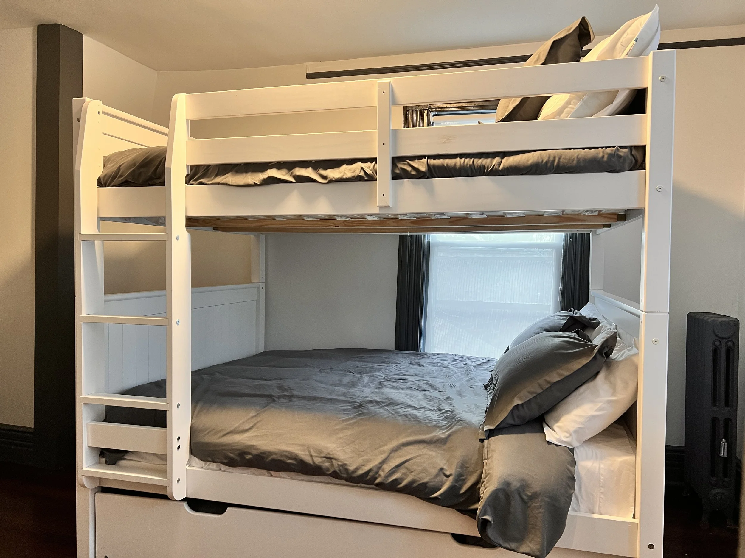White wooden bunk bed with a double bed on the bottom and a twin bed on top, against a beige wall with a window.