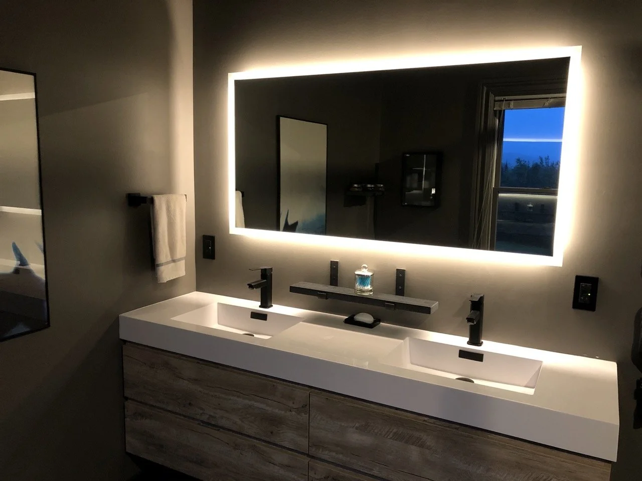 Modern bathroom vanity with two white sinks, black faucets, a large illuminated mirror, a towel hanger on the wall, and a window showing a blue sky with trees outside.