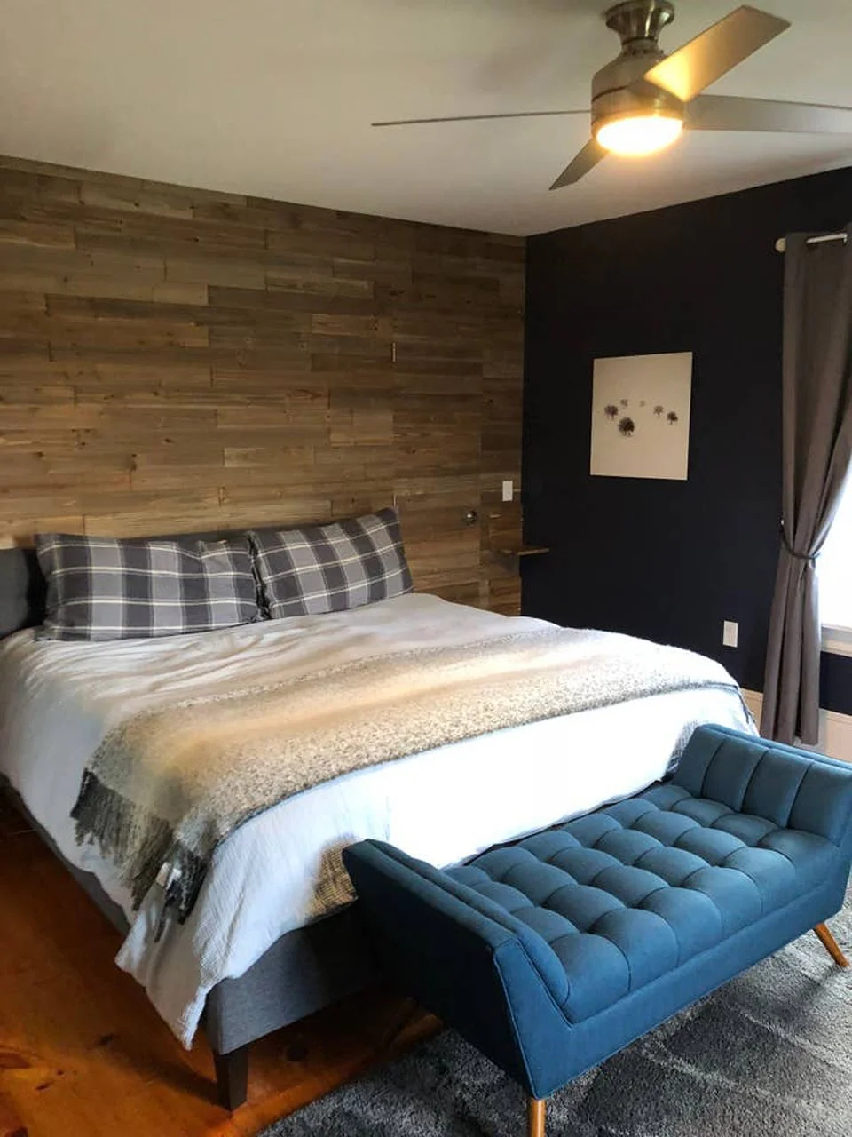 A bedroom with a bed having plaid pillows and a throw blanket, a wooden accent wall, a ceiling fan, a dark wall with a small wall art, curtains, and a teal upholstered bench at the foot of the bed.