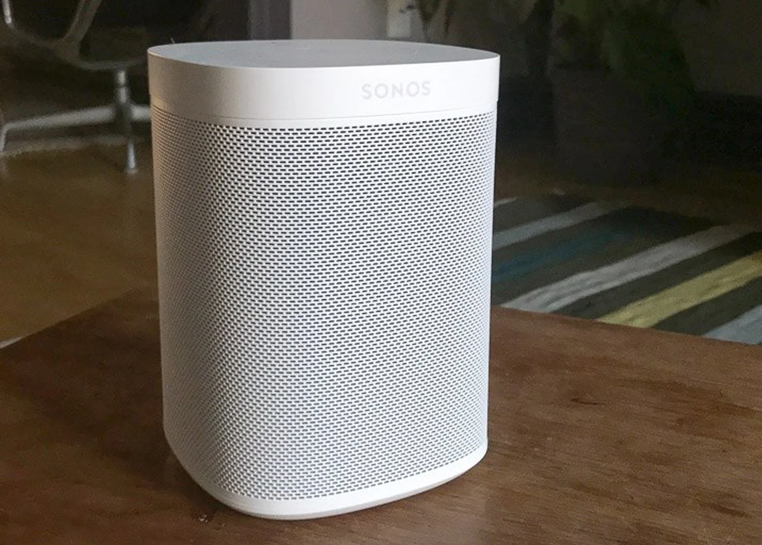A white Sonos speaker on a wooden surface with a couch and striped rug in the background.
