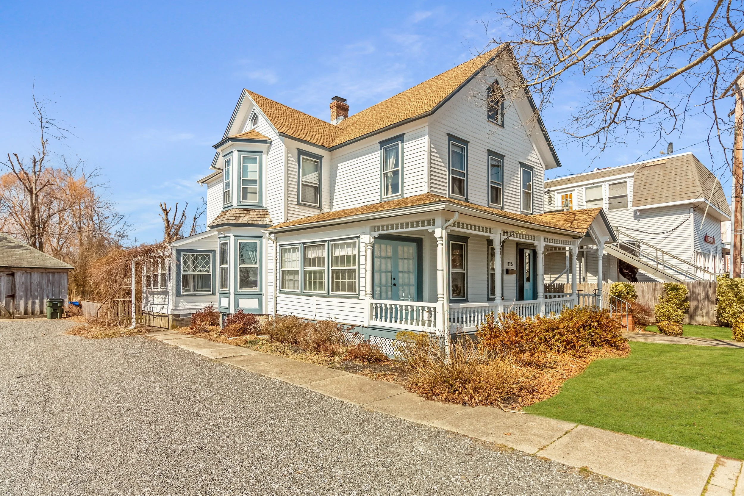 A large, white, Victorian-style house with multiple gabled roofs, bay windows, and a front porch with decorative trim, set against a blue sky with some clouds.
