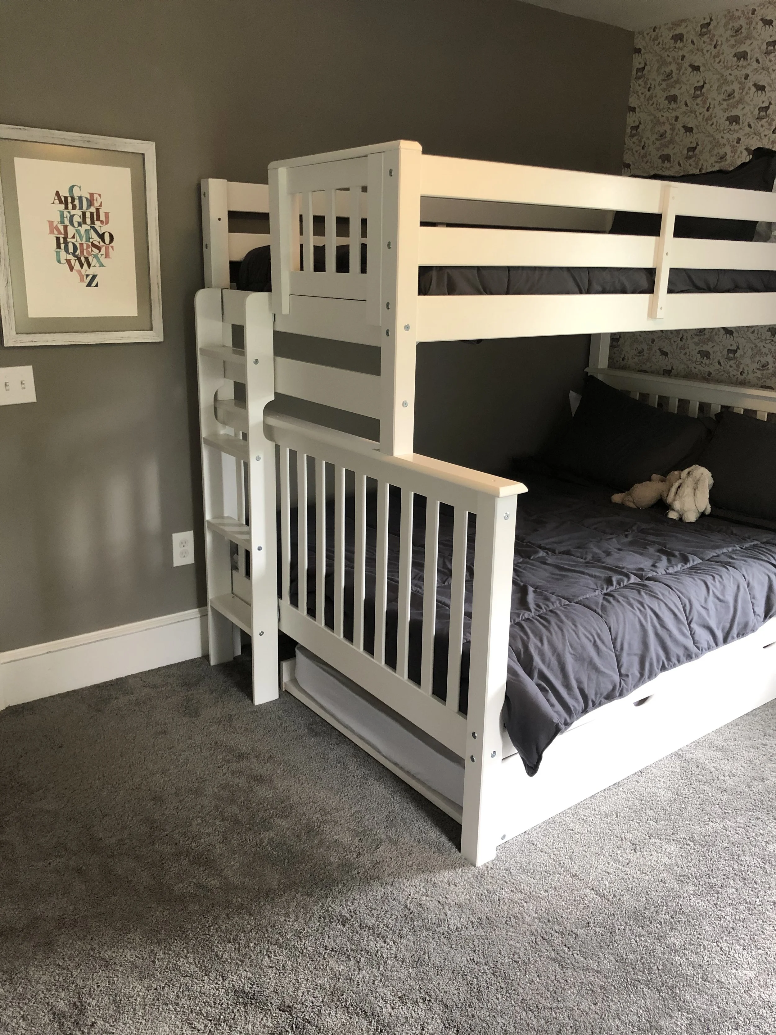 A white bunk bed with a ladder, set against a wall with gray paint and a patterned wallpaper with animals. The top bunk has a black mattress, and the bottom bunk has a black quilt with pillows, including a stuffed animal.