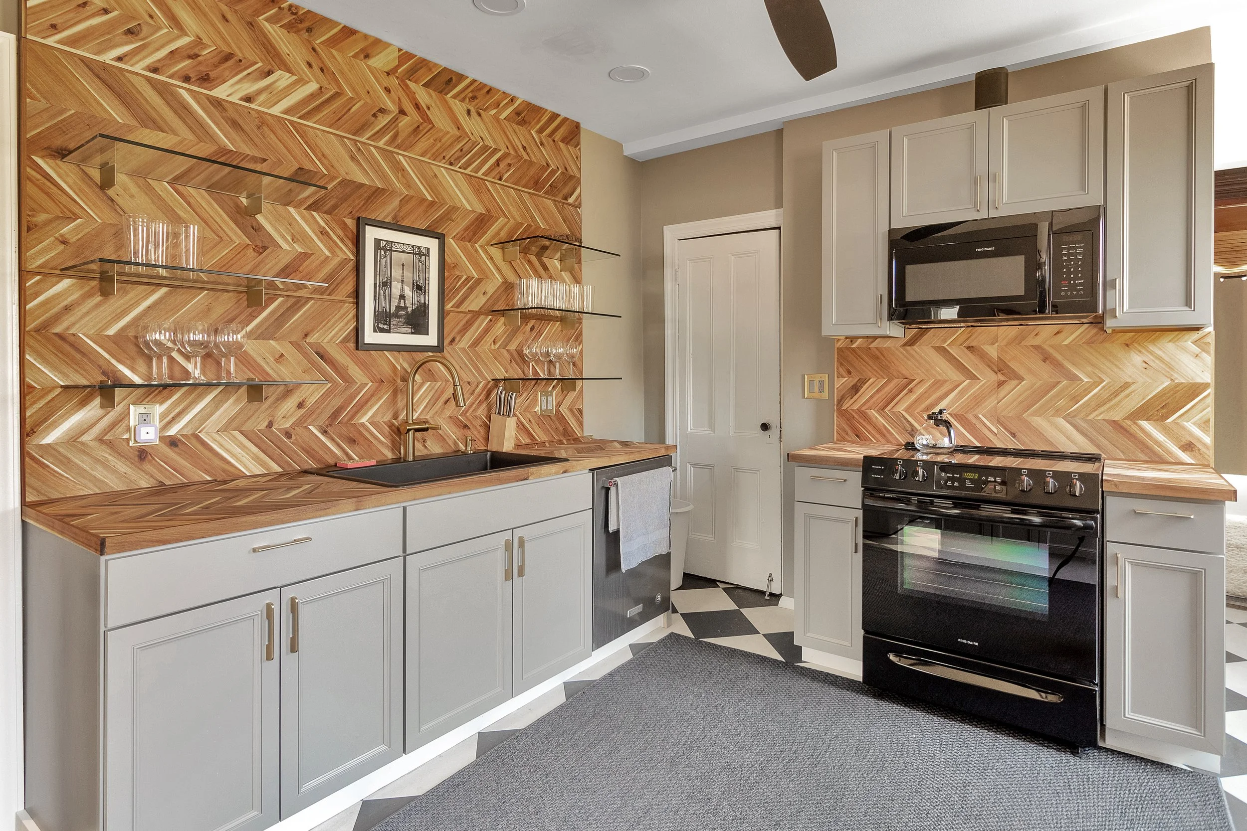 Kitchen with light gray cabinets, wooden countertops and backsplash, black stove, microwave, and small dishwasher, open shelving with glasses, and black and white checkered floor.