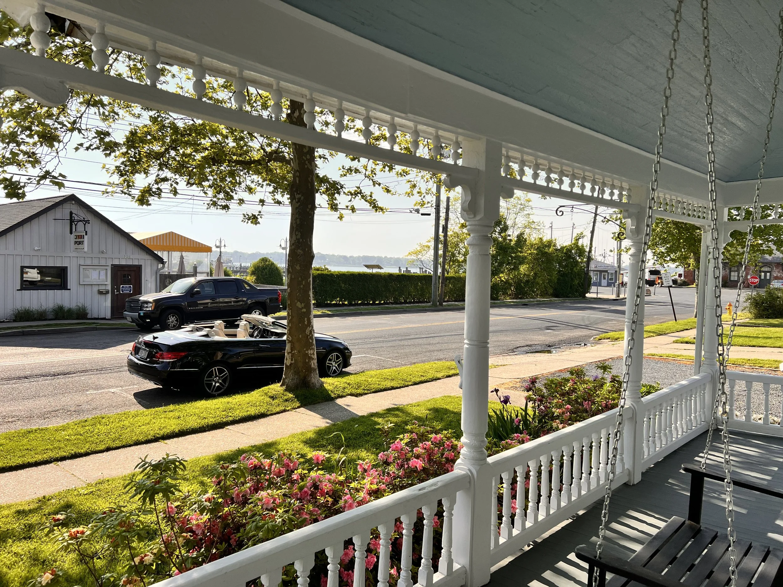View of a street from a porch with a white railing and floral garden, parked black convertible car, trees, and buildings in the background.