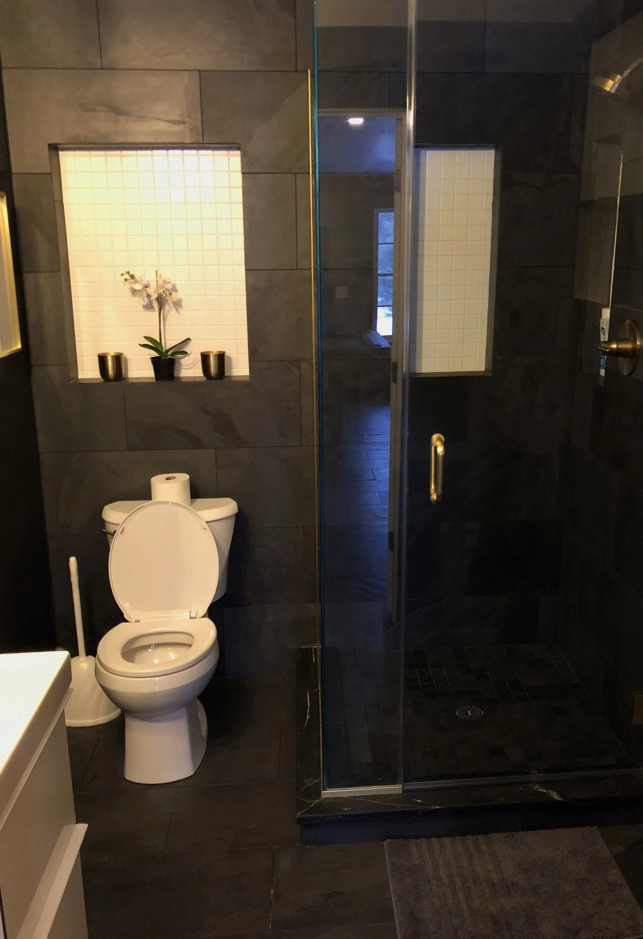 Bathroom with a toilet, a shower with dark tiles, and a small white tiled niche with an orchid and two small gold pots arranged inside.