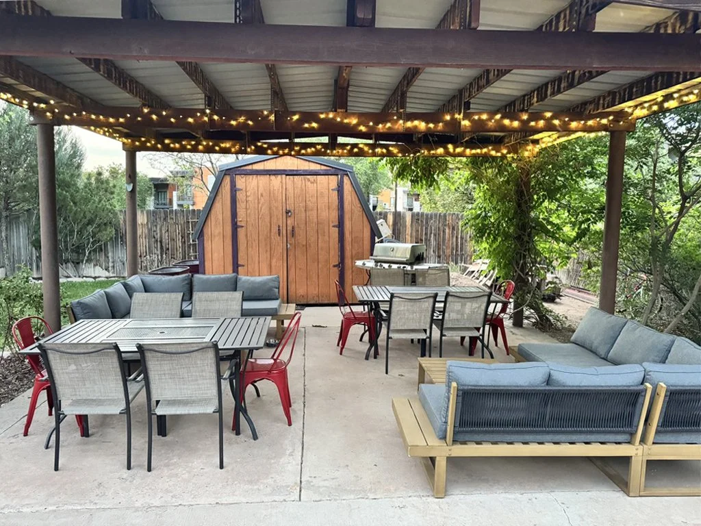 Covered outdoor patio area with string lights, outdoor sectional sofas, dining tables with chairs, a wooden shed, a barbecue grill, and surrounding greenery.