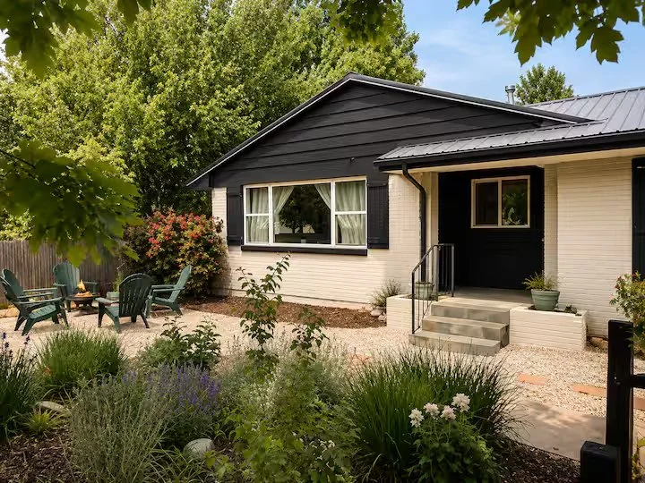 Front of a brick house with a porch, steps, and garden, surrounded by trees and outdoor chairs.
