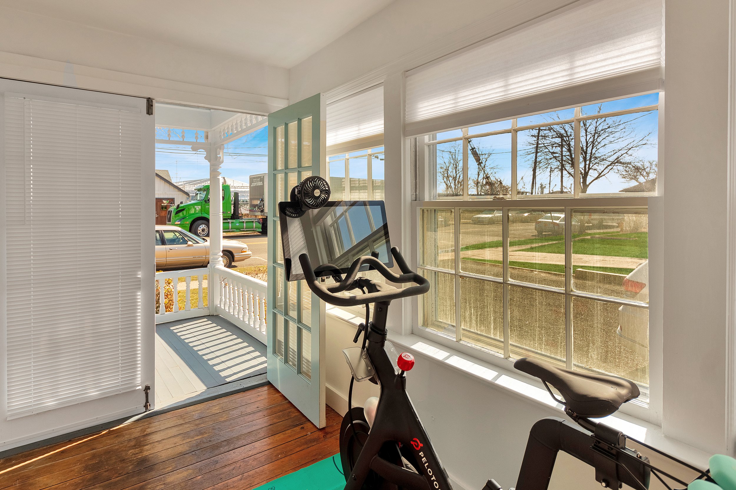 Indoor room with exercise bike facing large windows that look out onto a porch and street with cars and a large green truck.