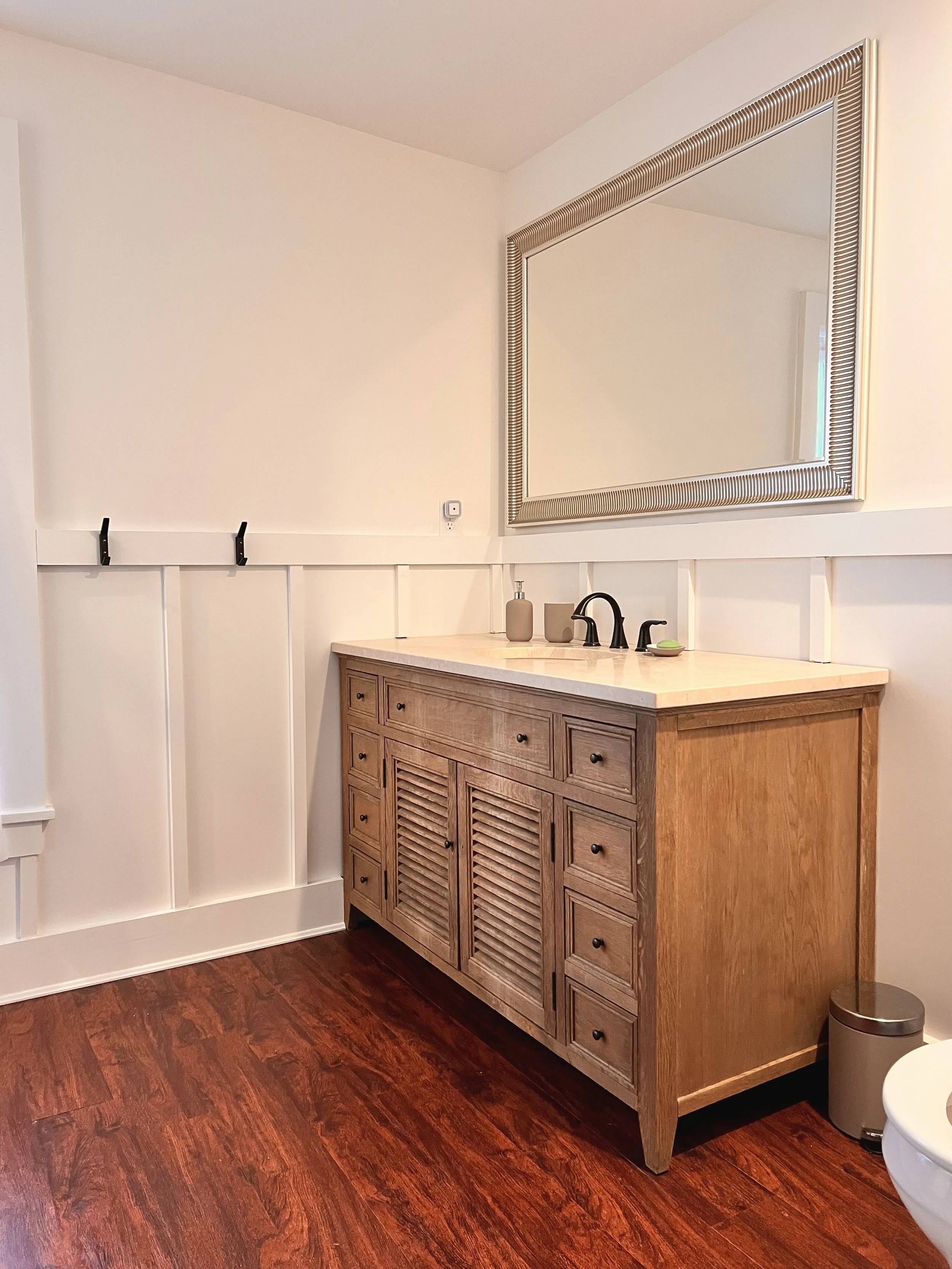 A bathroom vanity with a wooden cabinet, a white countertop, black faucet, and a large mirror above it. There are small decorative containers on the countertop and a trash bin next to the vanity. The wall behind has white paneling, and the floor is dark wood.