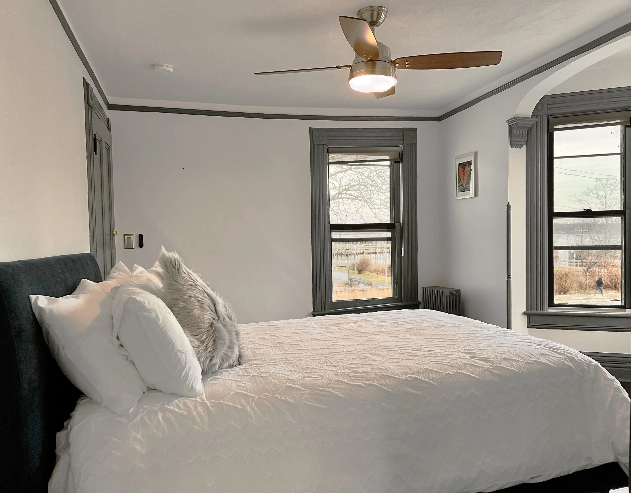 A cozy bedroom with a large bed covered in white bedding, gray headboard, fluffy pillow, and decorative fur pillow. The room features dark gray window frames, two windows showing an outdoor scene with trees and a person walking, and a ceiling fan with wooden blades.