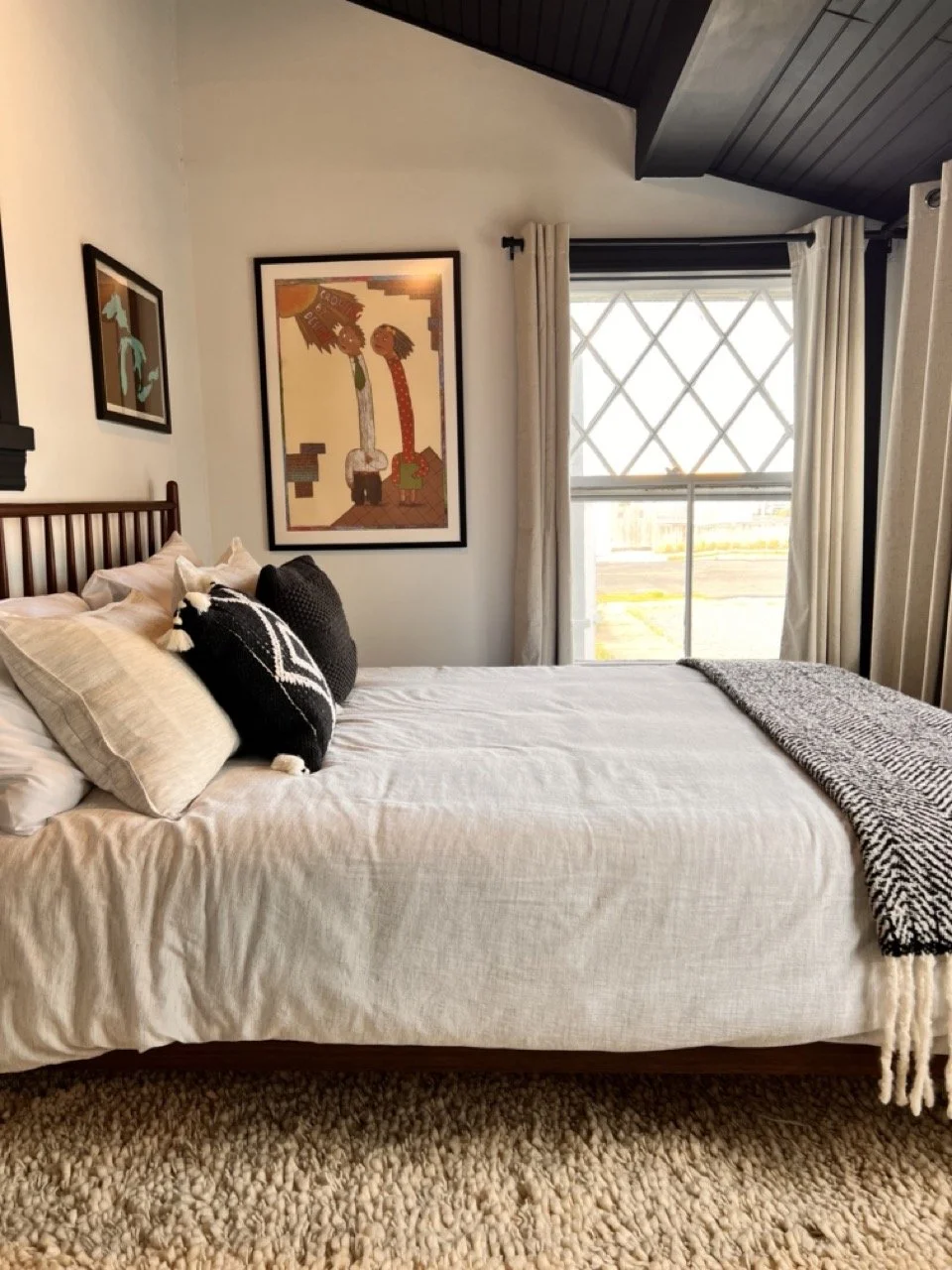 A cozy bedroom with a bed that has beige bedding and black-and-white decorative pillows, a textured throw blanket, a large window with diamond grid pattern, beige curtains, and artwork on the wall.