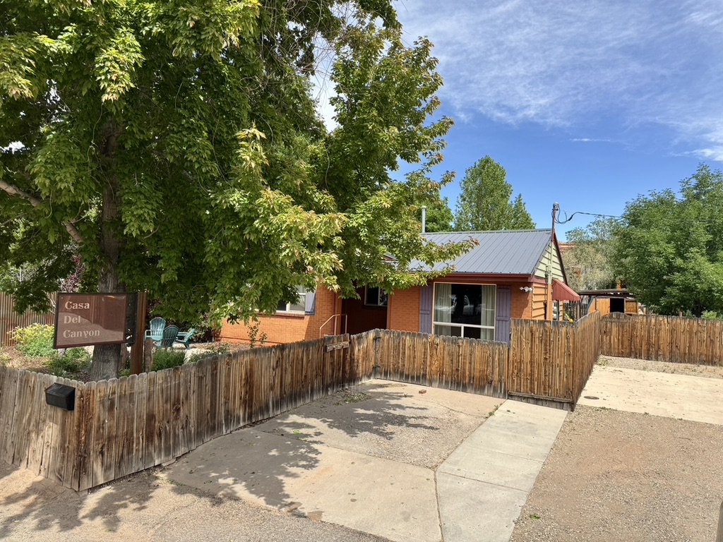 A small house with a metal roof, surrounded by a wooden fence, with a large green tree in front and a bright blue sky overhead.