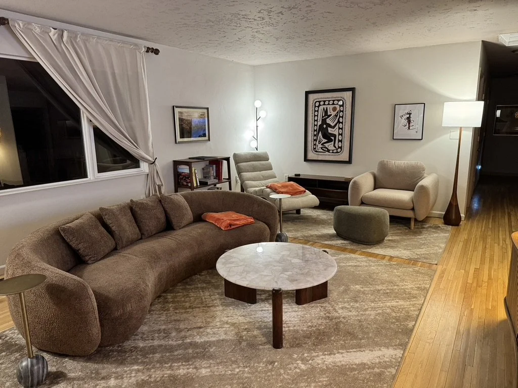 Living room with a curved brown sofa, a round marble coffee table, an off-white armchair, an adjustable lounge chair, a green ottoman, and a tall floor lamp. Wall art and a window with curtains are visible.