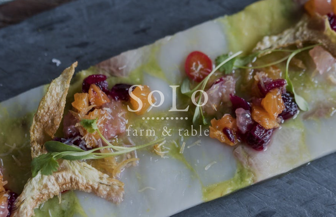Gourmet appetizer with diced fruits and microgreens on a crispy cracker, served on a dark slate board.