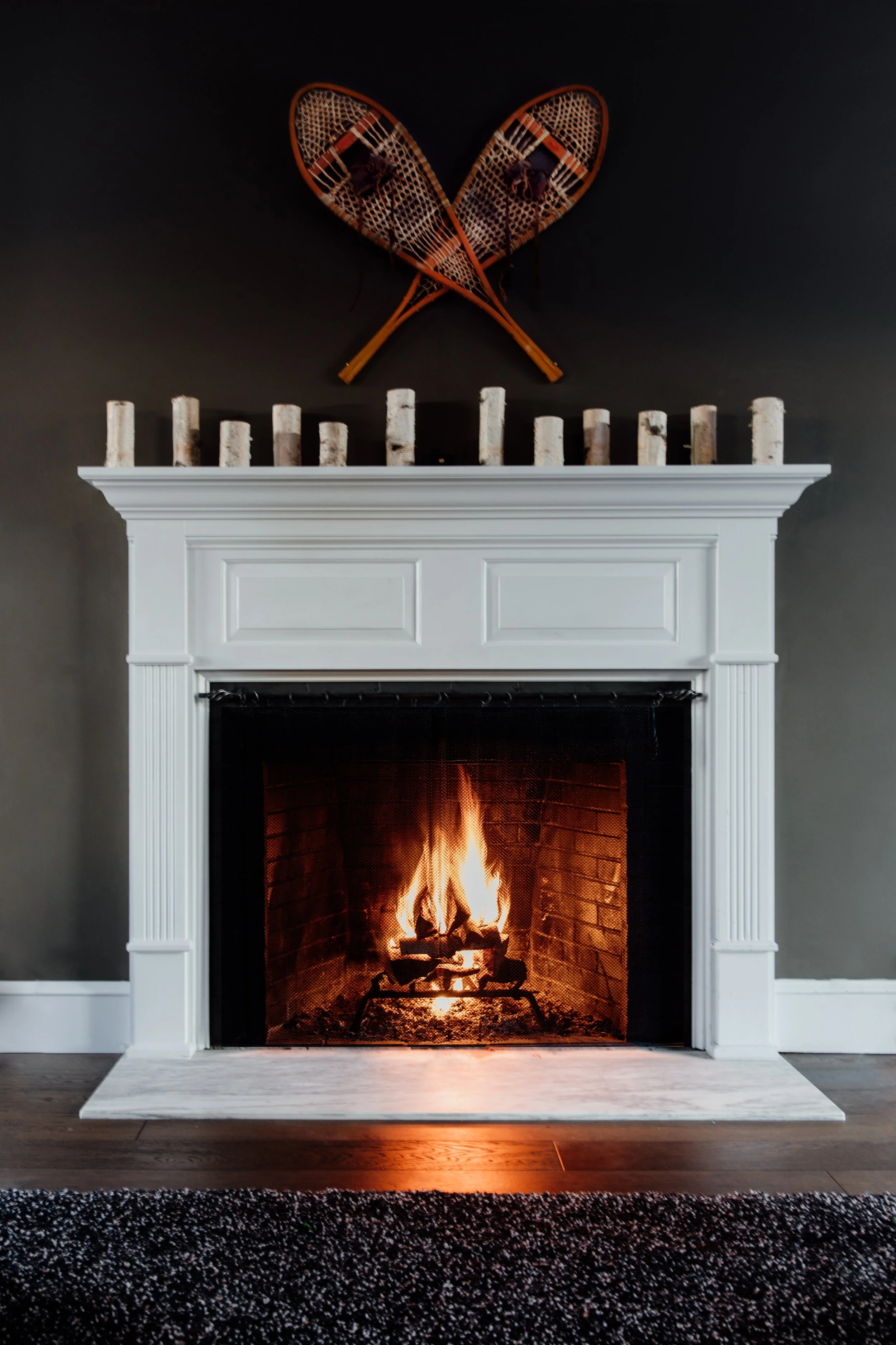A white fireplace with a fire burning inside, topped with a row of birch logs. Two vintage wooden snowshoes hang above the fireplace on a black wall.
