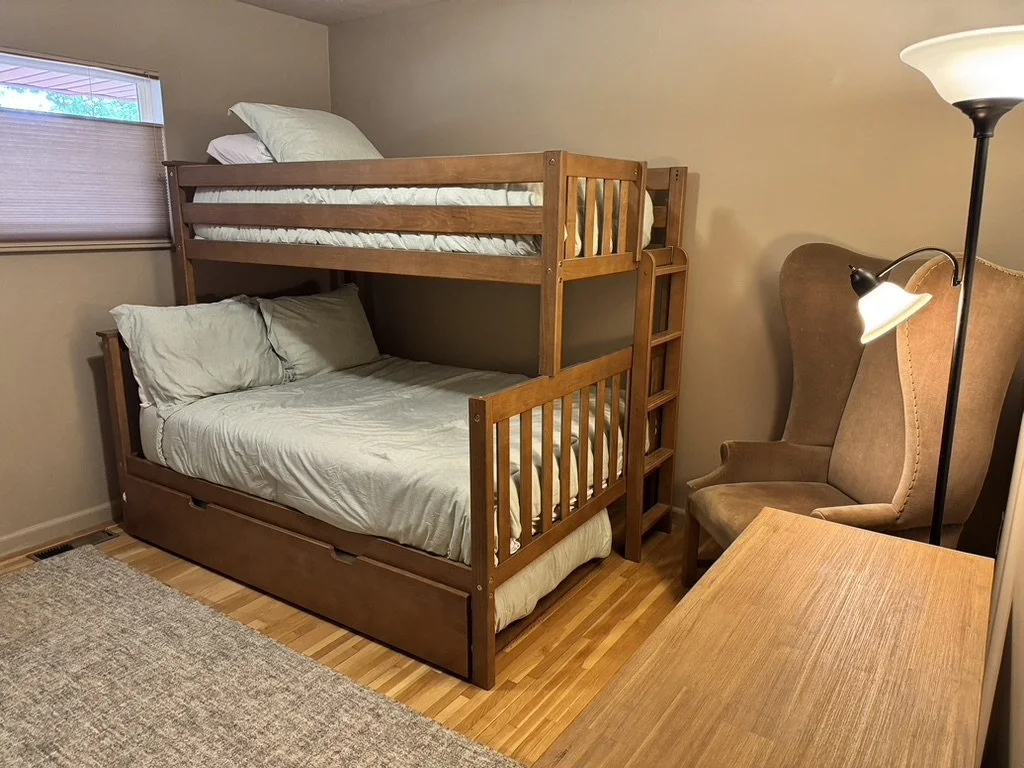 A bedroom with a wooden bunk bed, a beige wingback chair, a small wooden desk, a floor lamp, a window with closed blinds, and a beige area rug.
