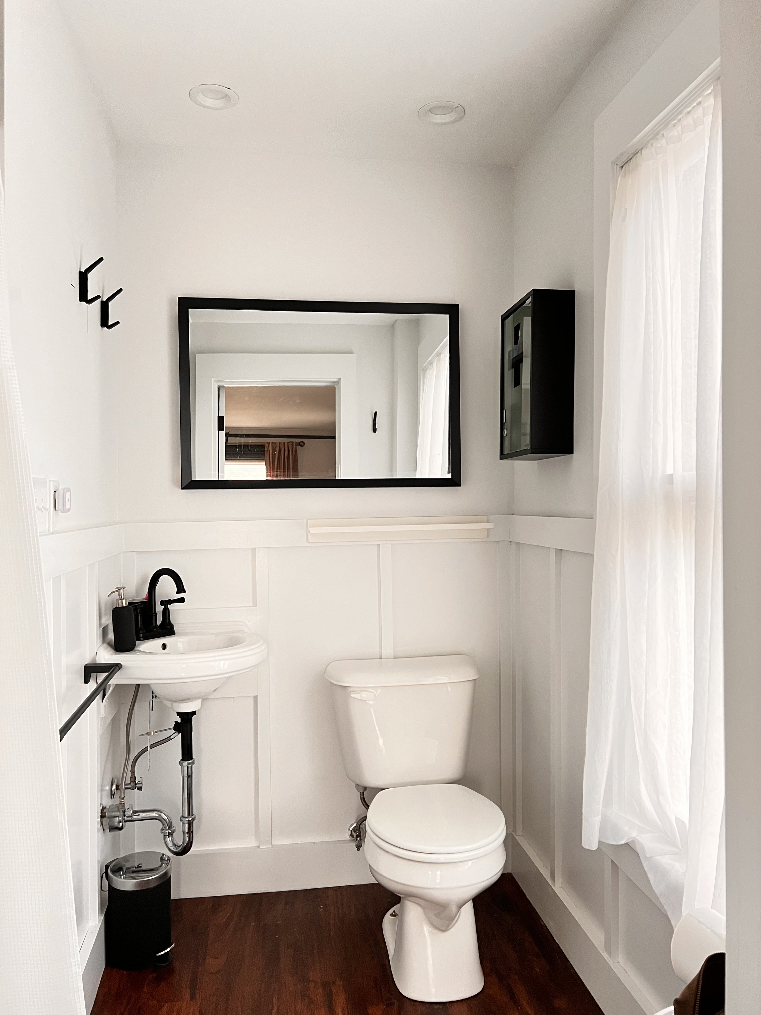 Small bathroom with white walls, a toilet, a tiny sink with black fixtures, a large mirror, and a window with white curtains.