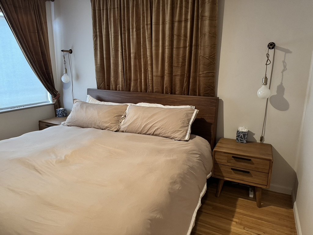A bedroom with a bed, beige bedding, and pillows, flanked by two wooden nightstands, each with a hanging pendant light. There's a window with brown curtains to the left.