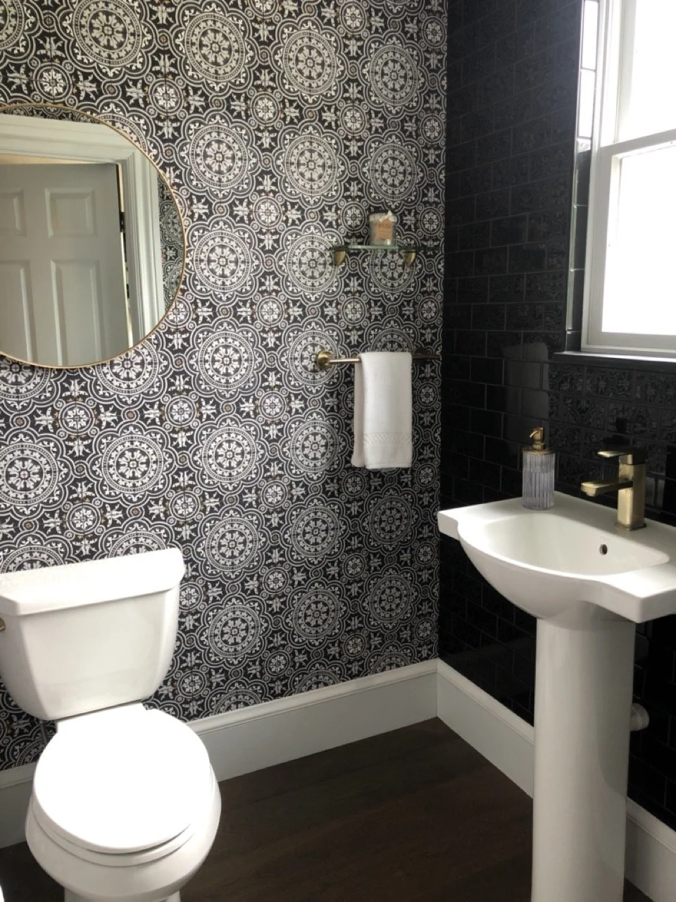 A small bathroom with patterned black and white wallpaper, a round mirror, a white toilet, a small pedestal sink with a gold faucet, a gold towel bar with a white towel, a glass soap dispenser, and a window letting in natural light.