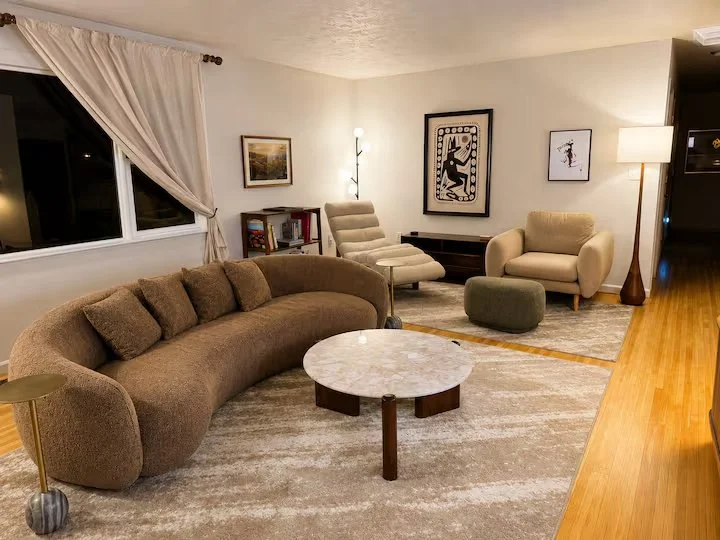 Living room with a curved brown sofa, a round marble coffee table, an off-white armchair, an adjustable lounge chair, a green ottoman, and a tall floor lamp. Wall art and a window with curtains are visible.