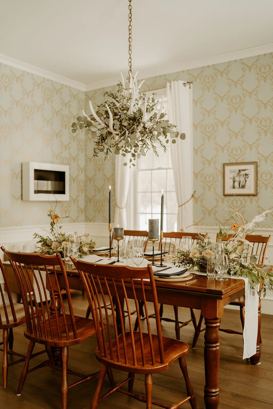 A dining room with a wooden table set for a meal, decorated with floral centerpieces, glassware, and black candles. The room has beige patterned wallpaper, white curtains, and a chandelier hanging above.