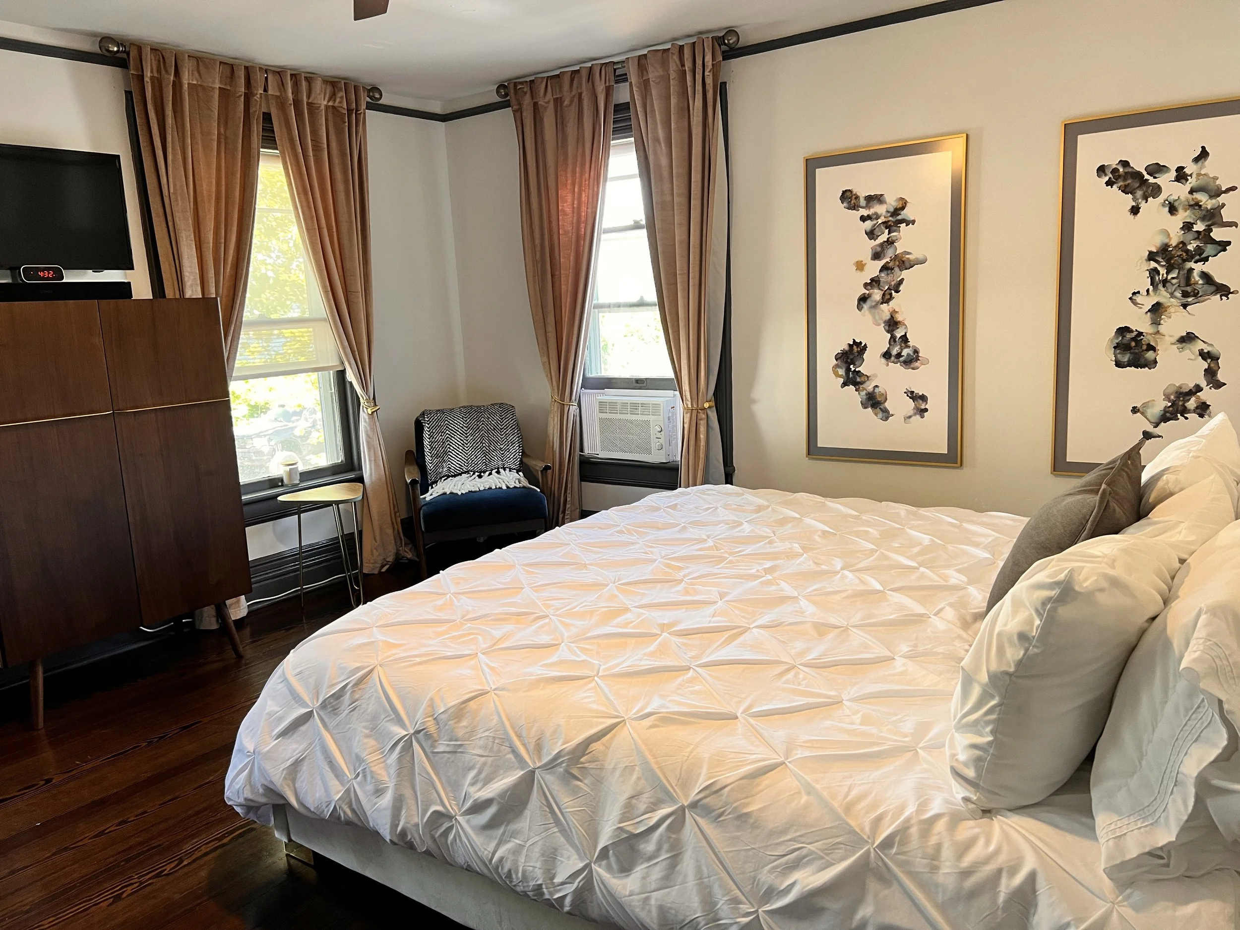 A cozy bedroom with two windows with beige curtains, a bed with white ruched bedding and gray pillows, a black armchair with a throw blanket, a small side table, a wooden cabinet with a TV on top, and two abstract framed artworks on the wall.