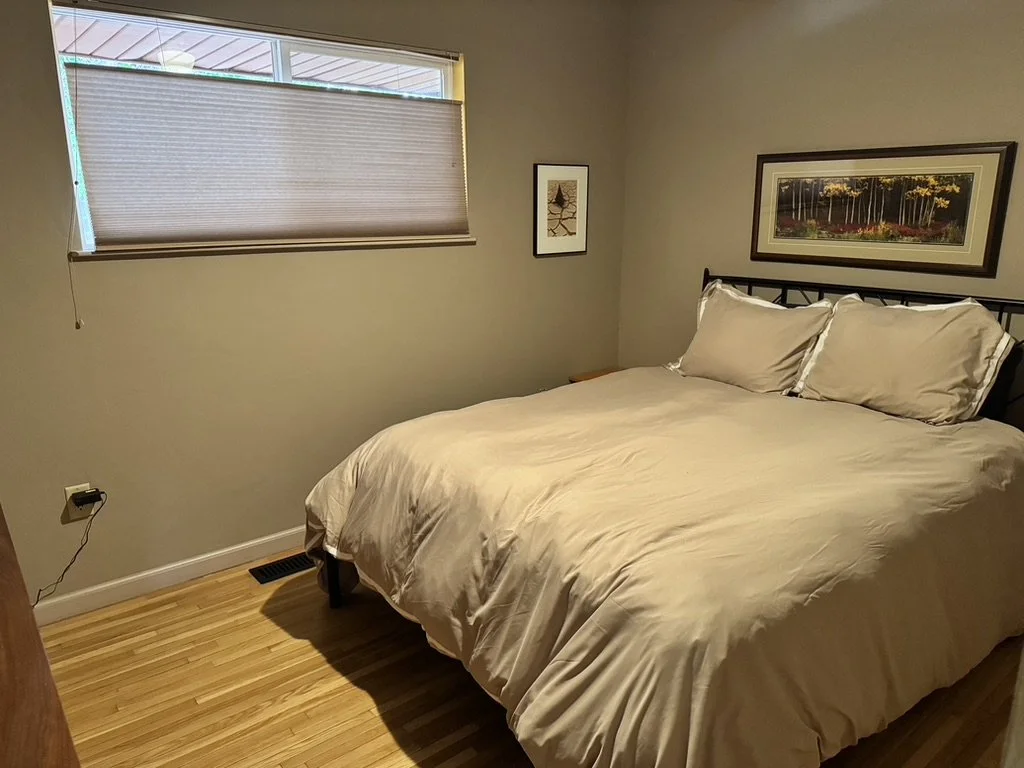 Small bedroom with a bed, beige bedding, pillows, window with a closed beige shade, and framed wall art.