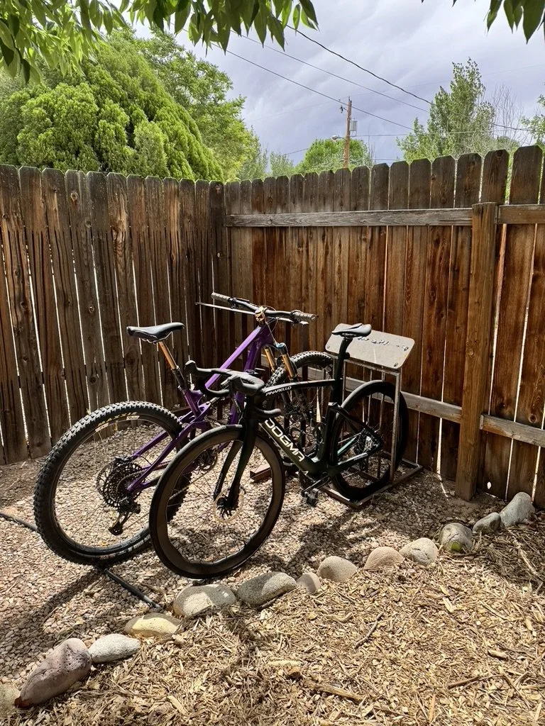 A backyard with a wooden fence, two bicycles (one mountain bike and one e-bike), and a small gravel area enclosed by a border of rocks. Overcast sky with some trees and power lines in the background.