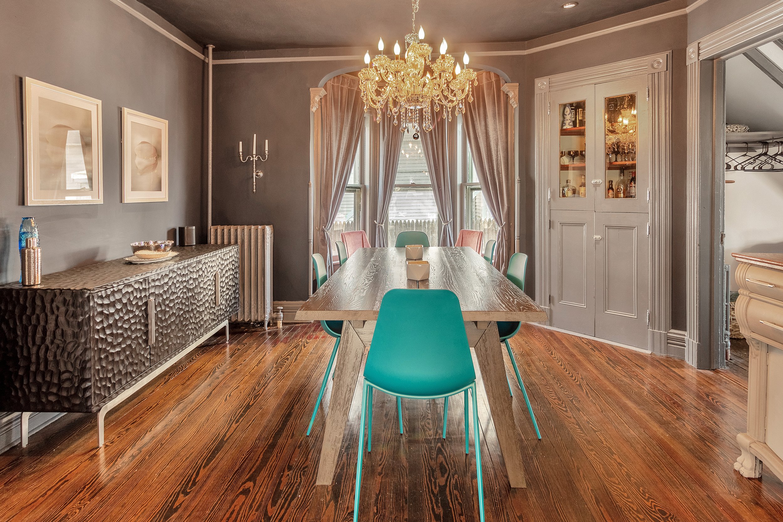 A dining room with a wooden table surrounded by teal chairs, a chandelier hanging from the ceiling, and windows with beige curtains in the background. There is a sideboard on the left and a glass-front cabinet on the right.
