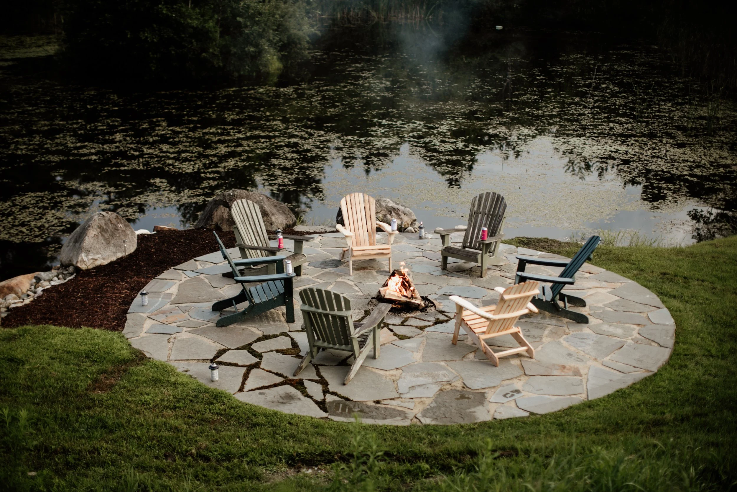 Outdoor fire pit area with eight Adirondack chairs arranged in a circle on a stone patio near a pond.