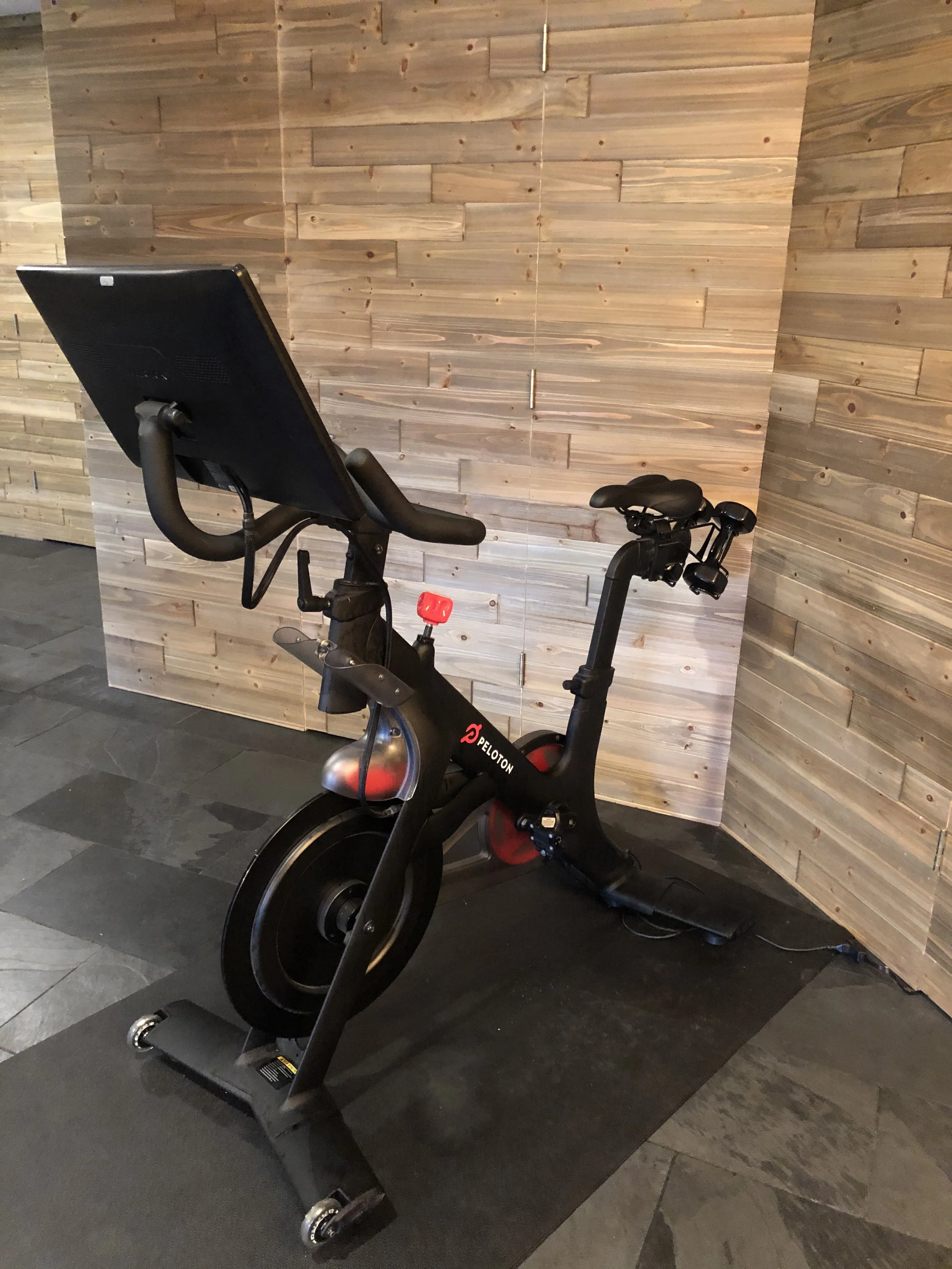 A Peloton stationary exercise bike with a screen mounted on the handlebars, situated in a room with wooden walls and dark tile flooring.