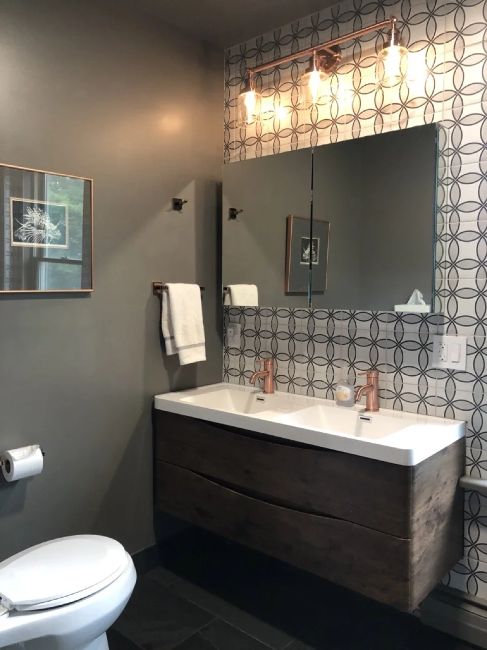 Modern bathroom with double sinks, rose gold fixtures, patterned tile wall, wall-mounted mirror, artwork, copper light fixtures, and a toilet.