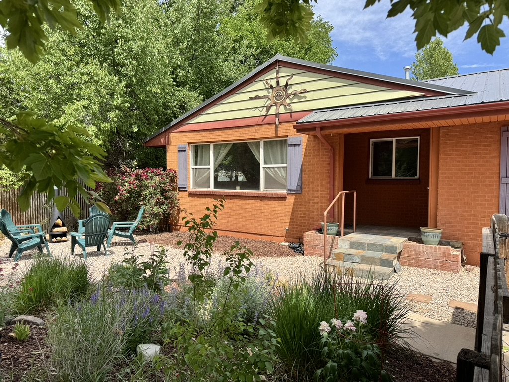 Front of a brick house with a porch, steps, and garden, surrounded by trees and outdoor chairs.