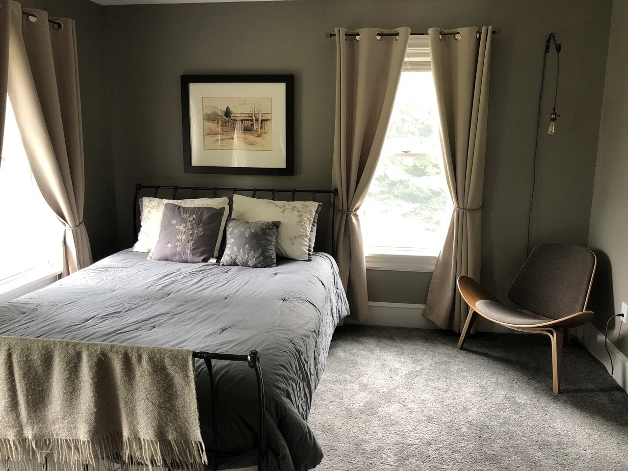A cozy bedroom with a bed covered in a gray duvet, four decorative pillows, beige curtains on windows, a framed landscape painting above the bed, a modern chair in the corner, and a light bulb hanging from the ceiling.
